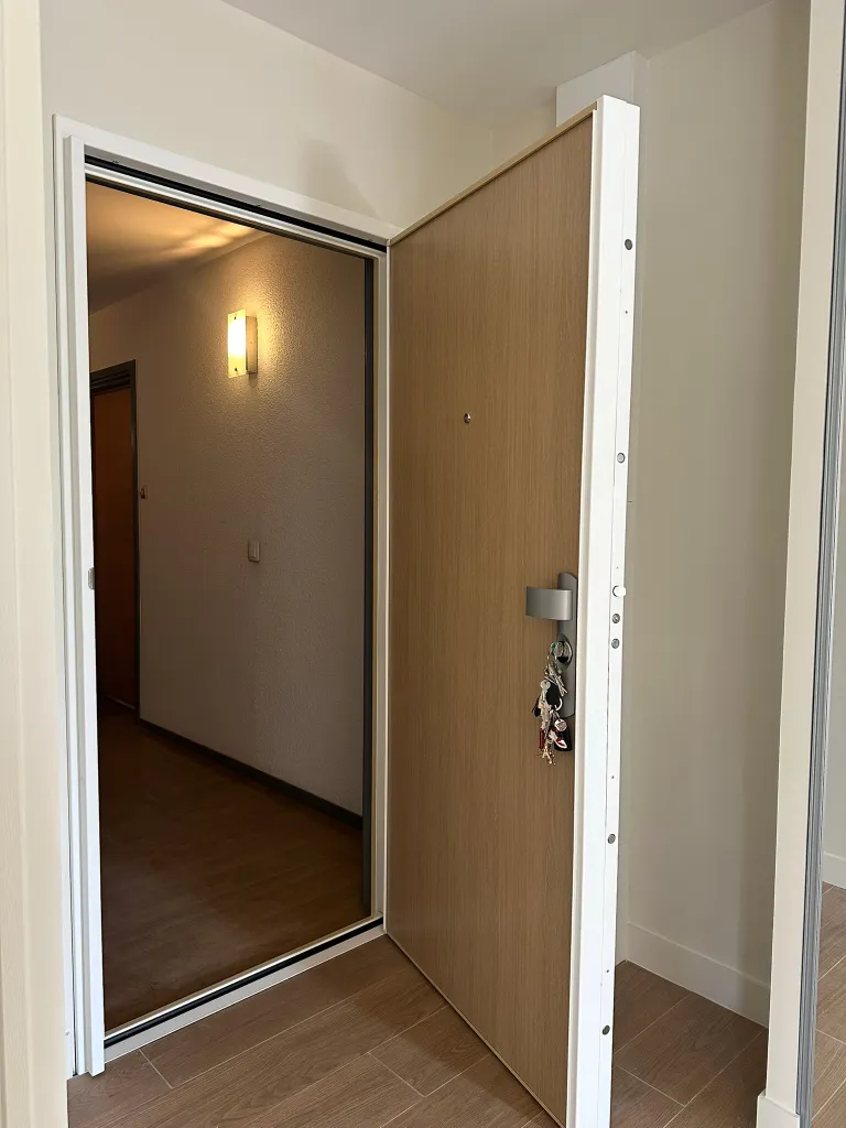 Bloc-porte blindé finition bois clair, ouvrant entrebâillé laissant voir le chant en acier et les pênes du système multipoints, posé sur un palier d'immeuble parisien